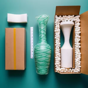 Illustration of packaging a glass vase for shipping. Steps include choosing a sturdy box slightly larger than the vase, wrapping the vase in bubble wrap and securing it with tape, filling the bottom of the box with packing peanuts, placing the wrapped vase inside, adding more packing peanuts to fill any remaining space, supporting the neck and base of the vase with molded foam or cardboard, and labeling the box as 'Fragile' and 'This Side Up'.